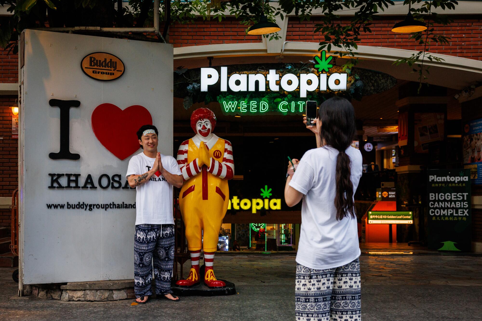 A man holds his palms together next to a statue of a clown as a woman takes his picture with a phone 