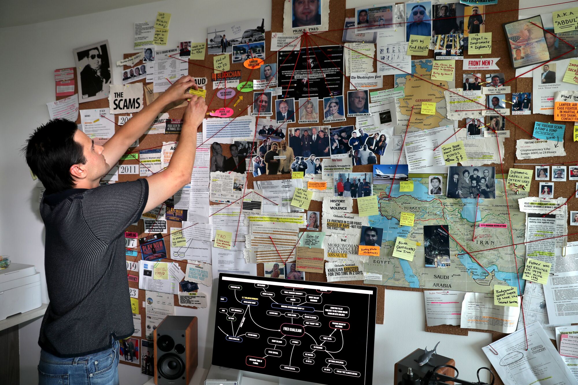 A man at left stands near a large cork board full of news clippings, notes and photos. He's pinning something up