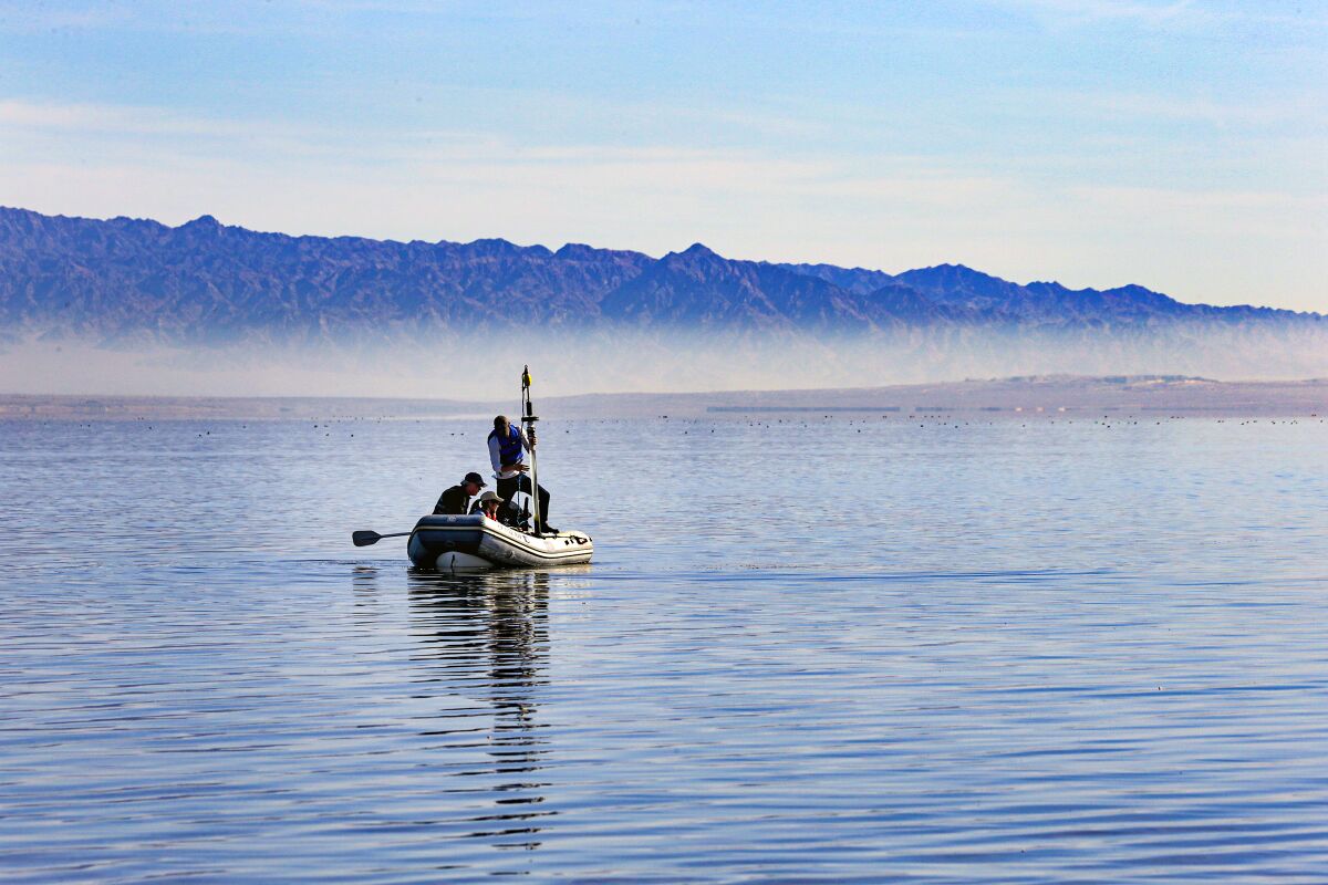 Three people sit in an inflatable craft on a body of water. In the background are low mountains.