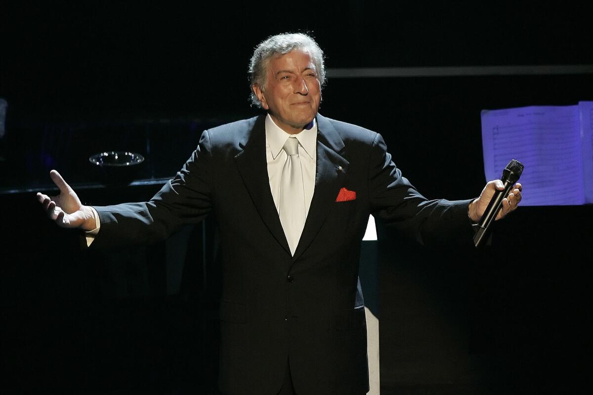 Tony Bennett holds his arms open as he wears a black blazer with a white shirt and tie while performing onstage 