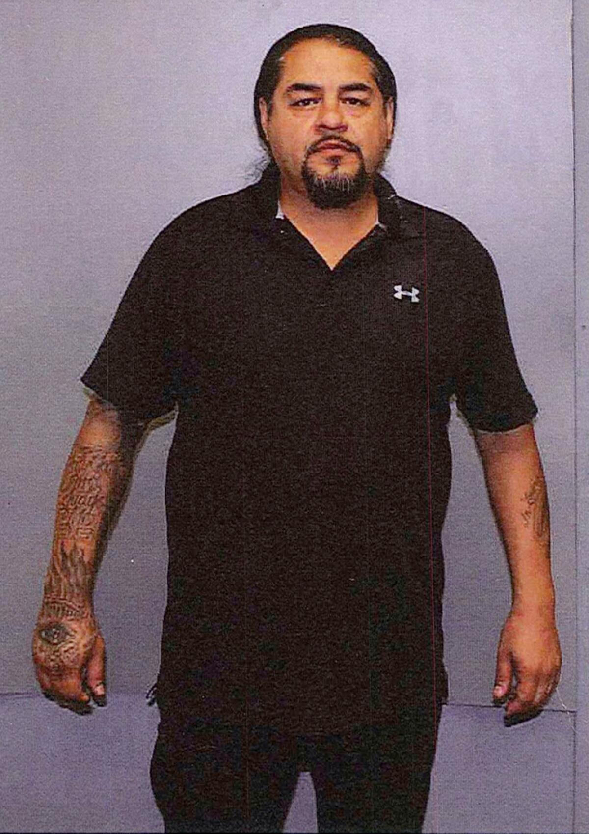  A man in work clothes with tattoos on his forearms stands before a blank background.