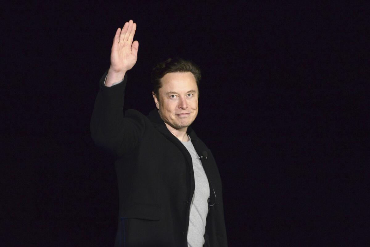 A man dressed in a light grey shirt and a black jacket waves with one hand toward the camera.