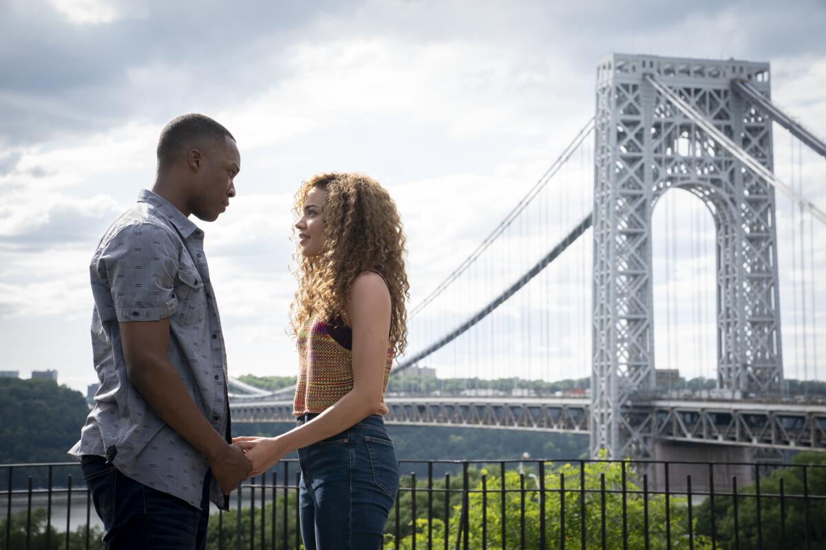 Corey Hawkins and Leslie Grace clasp hands in front of a bridge in the movie "In the Heights."