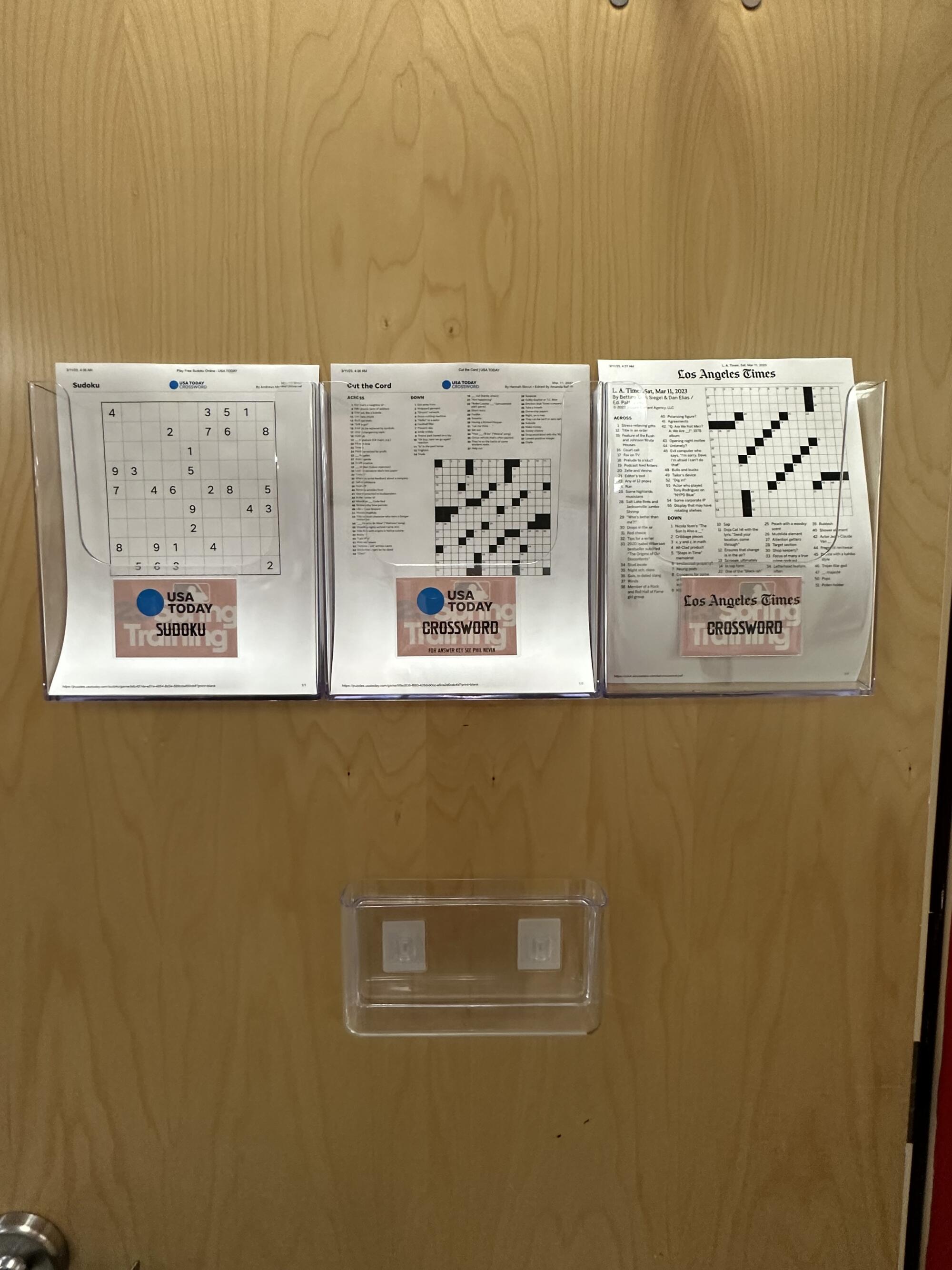 Sudoku and crossword puzzle printouts in clear bins on a wooden door.