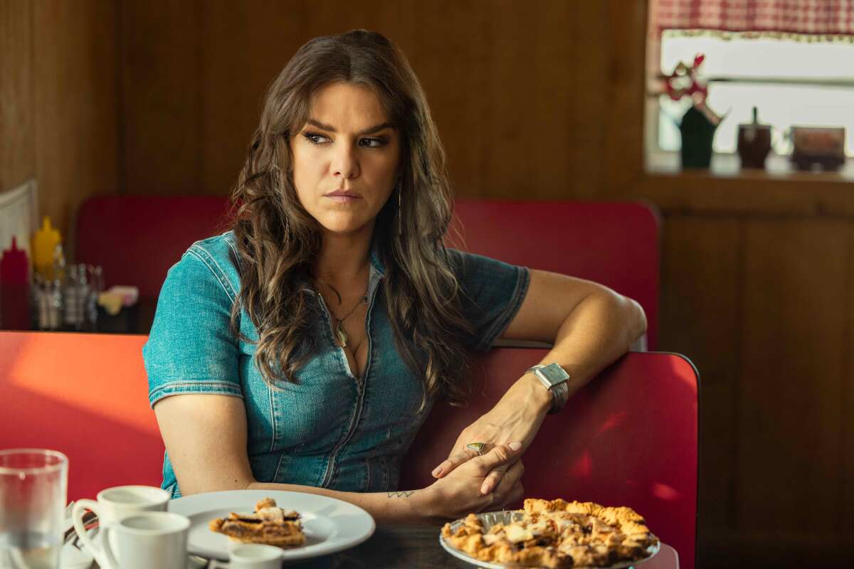 Uma mulher com blusa jeans e longos cabelos castanhos está sentada em uma mesa com uma fatia de torta na frente dela e olha para a direita.