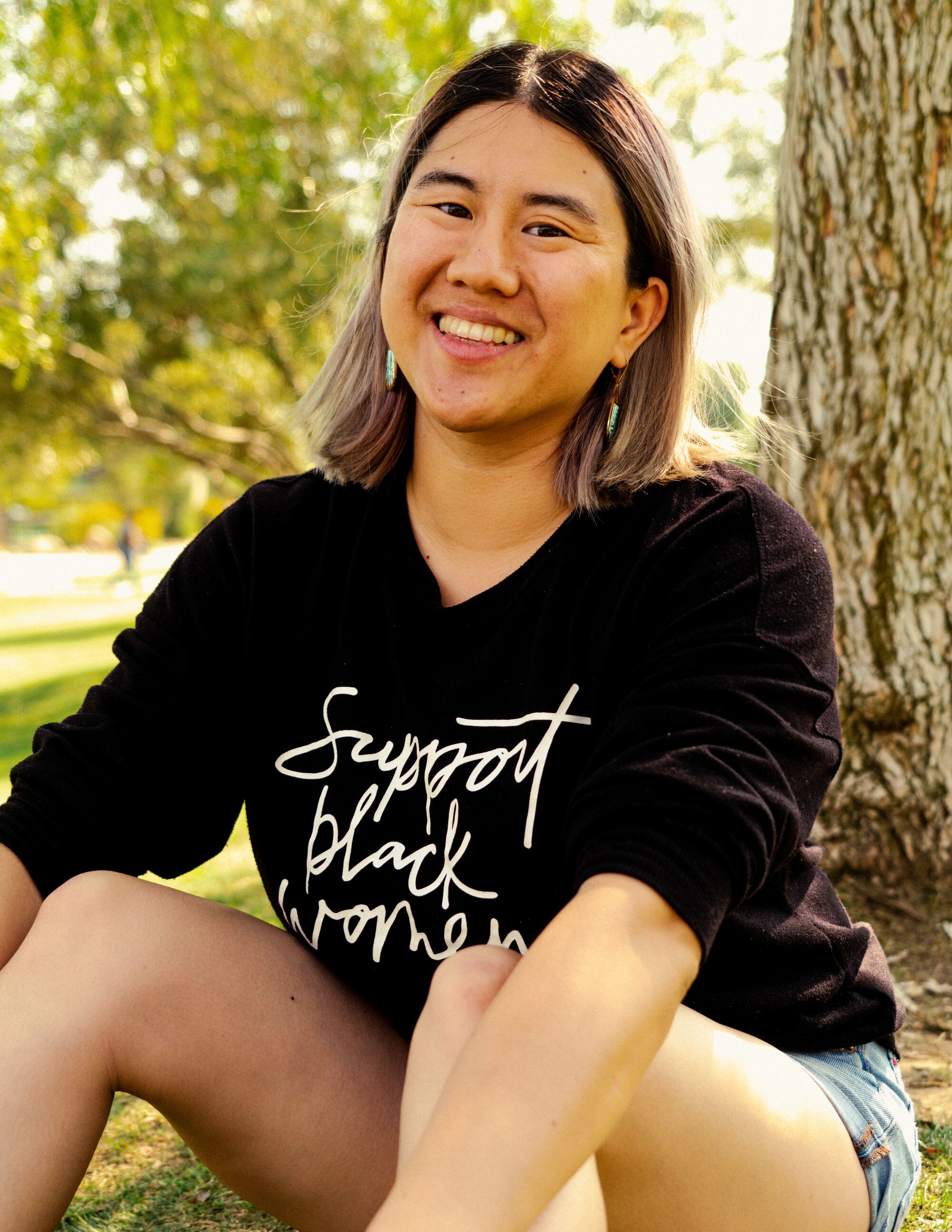 A person seated outdoors, wearing a sweatshirt that says "Support black women" and shorts.