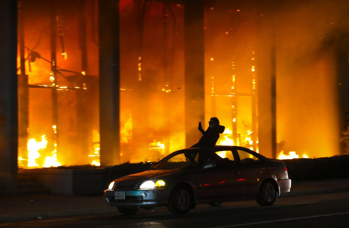 A person stands out of the sunroof of a car driving past a burning building