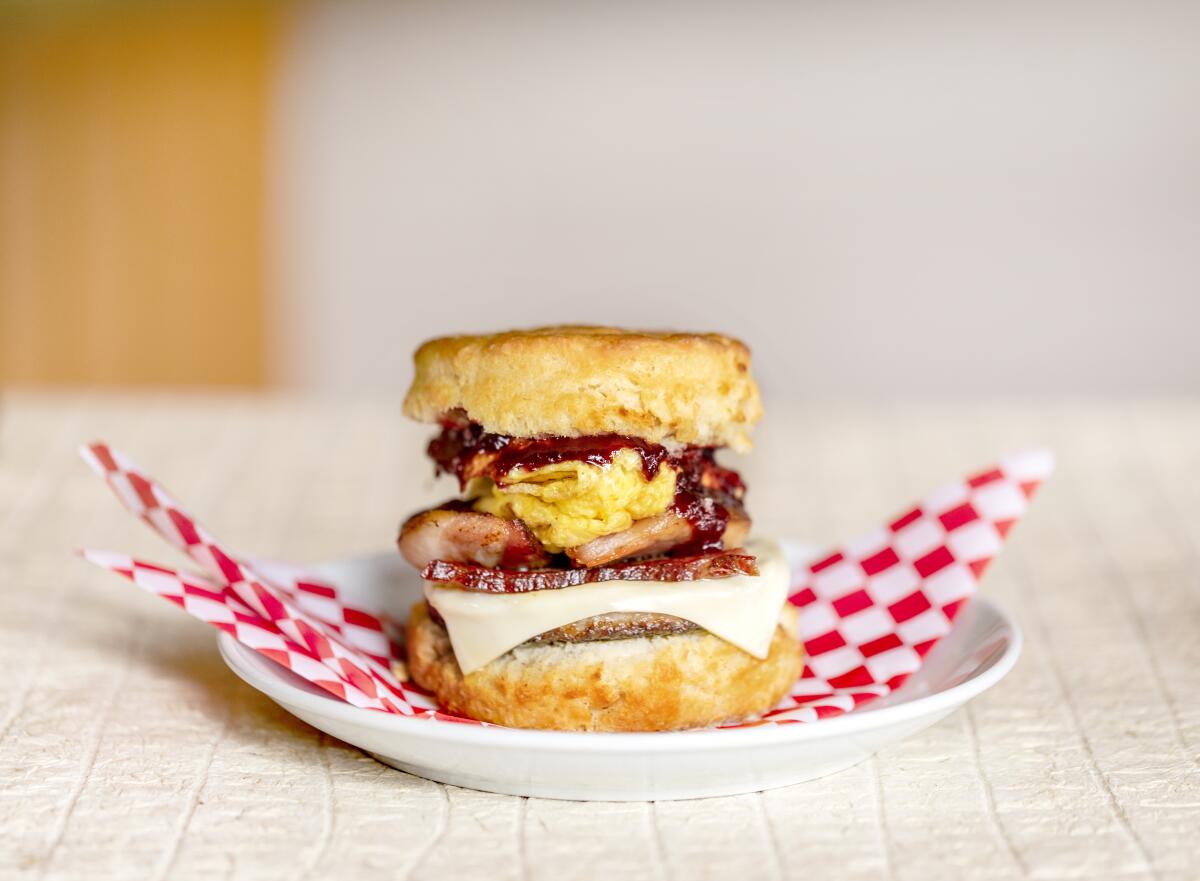 Closeup of egg, meats and cheese on a biscuit. The sandwich is on a white plate with a red and white checked napkin.