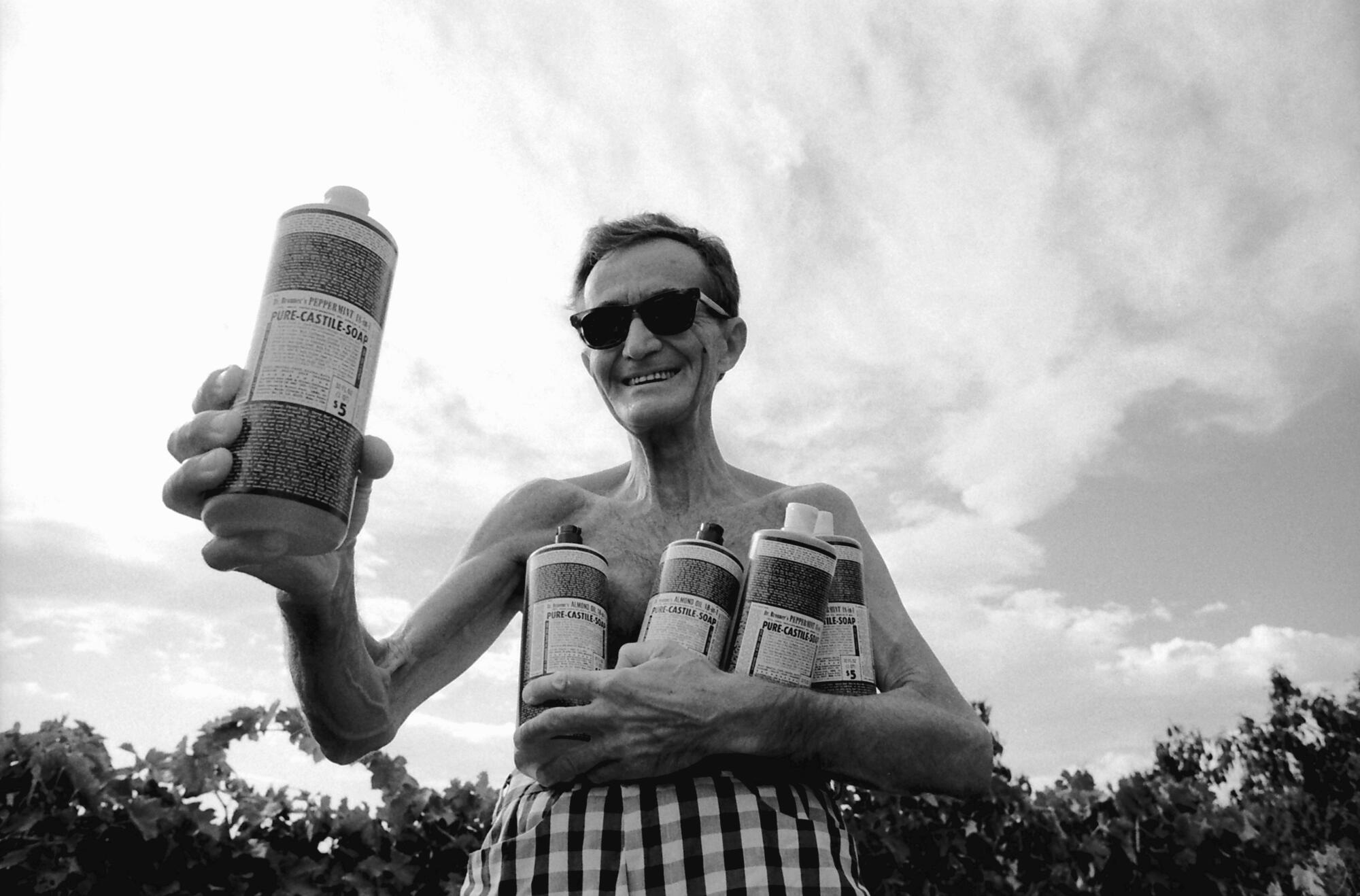 A black-and-white photo of a shirtless man outdoors, raising a labeled bottle of liquid and cradling several more in his arm.