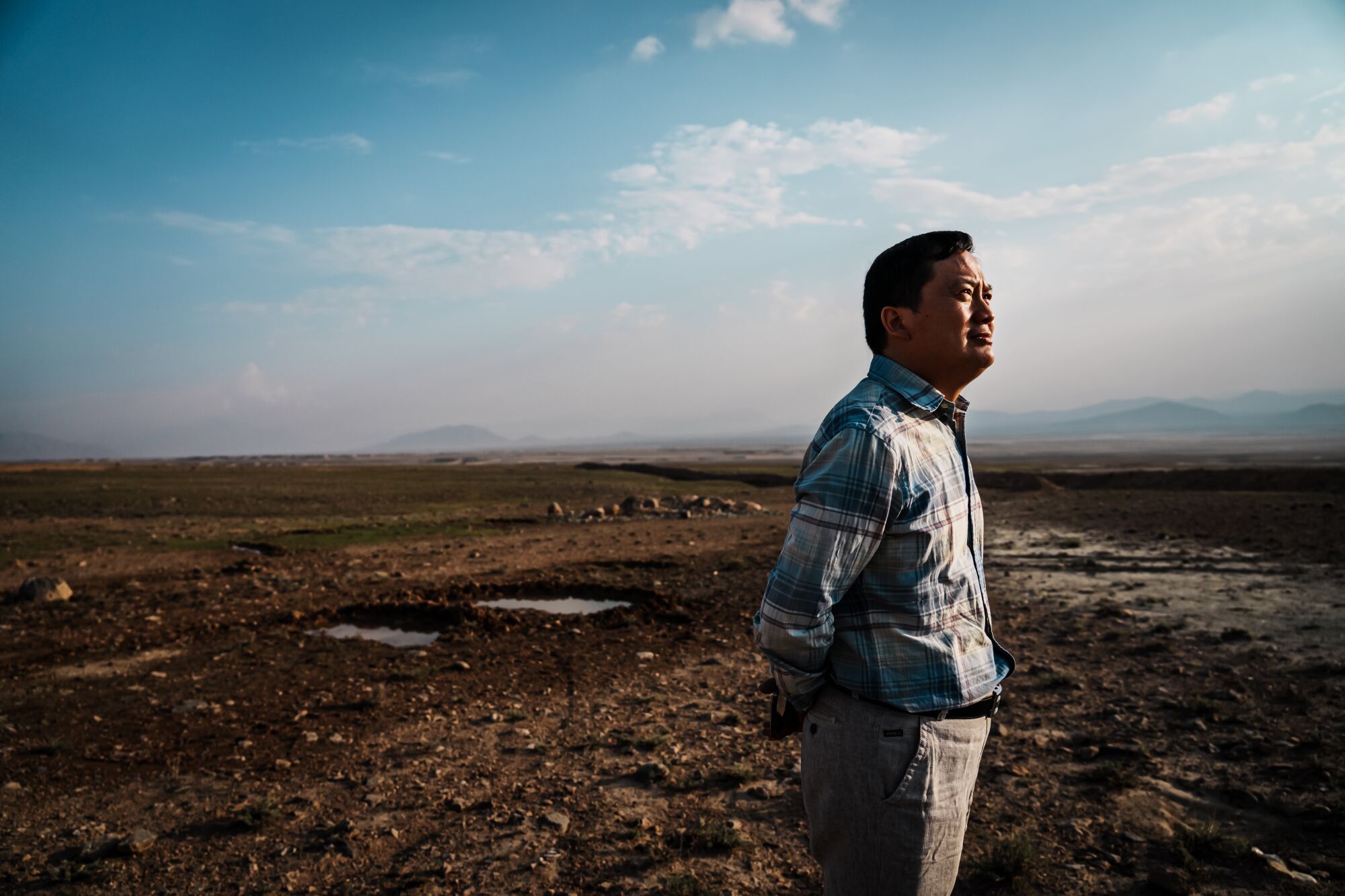 A man in a blue checked shirt and khaki pants stands in the middle of a barren land under a blue sky
