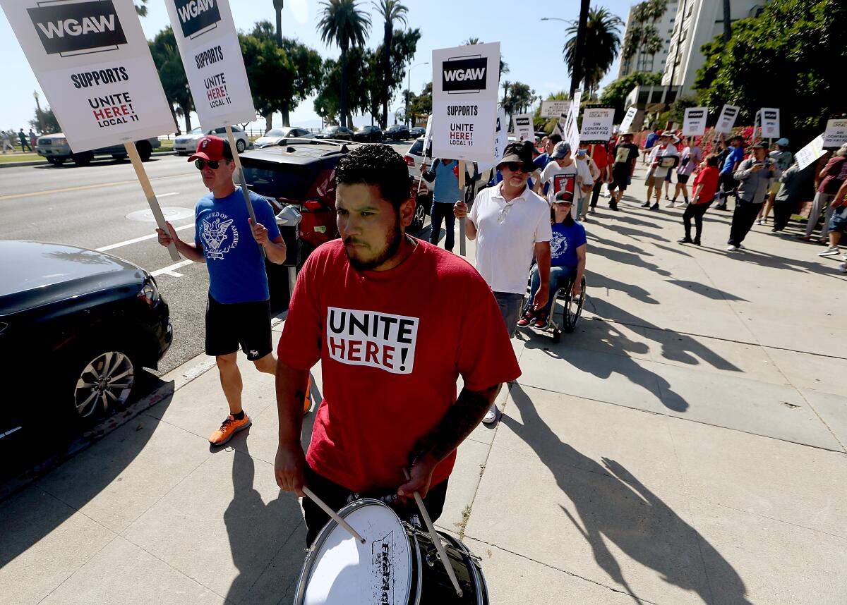 People hold signs and picket, one in a red "Unite Here" shirt, on a sidewalk with palm trees in the background