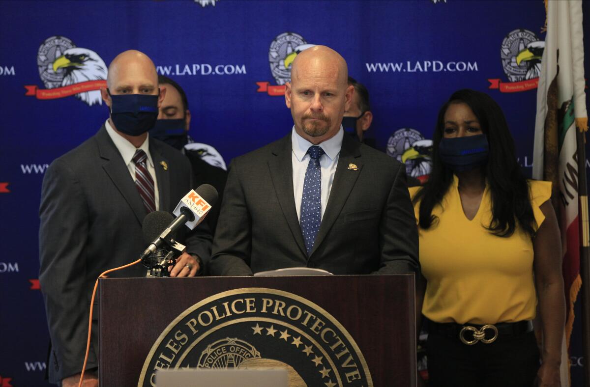 Jamie McBride speaks from behind an LAPD Protective League logo as others stand by against a blue LAPD.com eagle backdrop