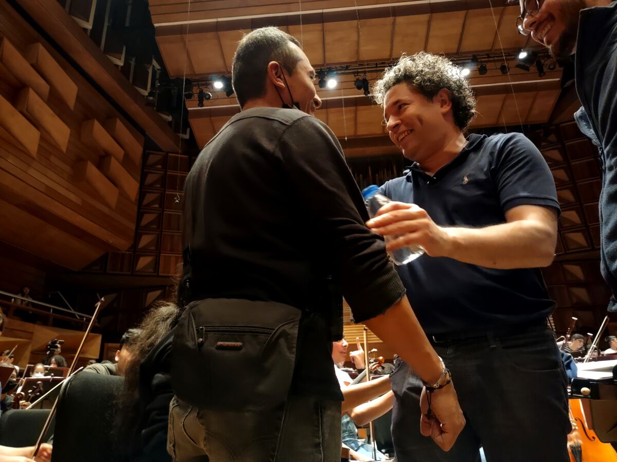 Gustavo Dudamel is seen about to embrace a young man in a rehearsal room with an orchestra seated behind him.