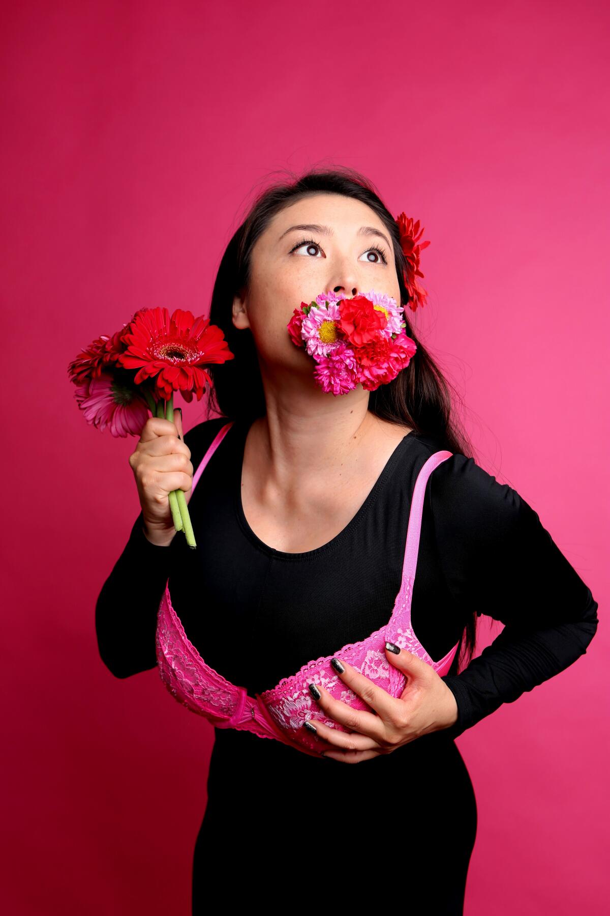 A woman with flowers in her mouth and hand wears a pink bra over an all-black outfit.
