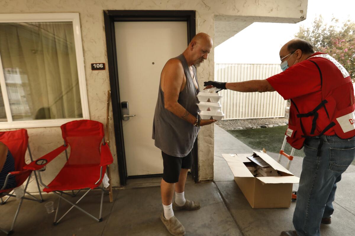 A Red Cross volunteer in a mask hands foam boxes of meals to a fire evacuee outside a motel room