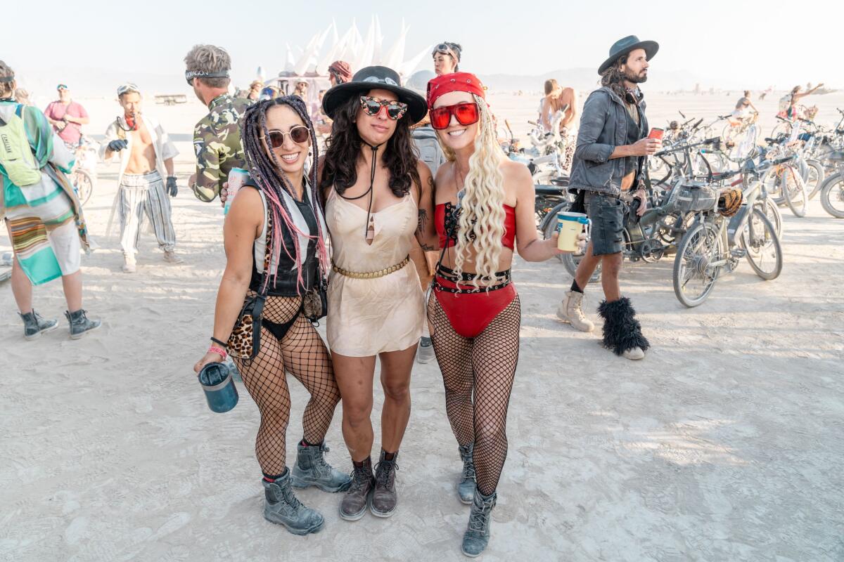 Three people posing on the playa at Burning Man while other people walk behind them.