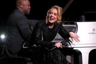 Adele wears a black beaded gown and leans over a white piano seat as a piano player plays behind her