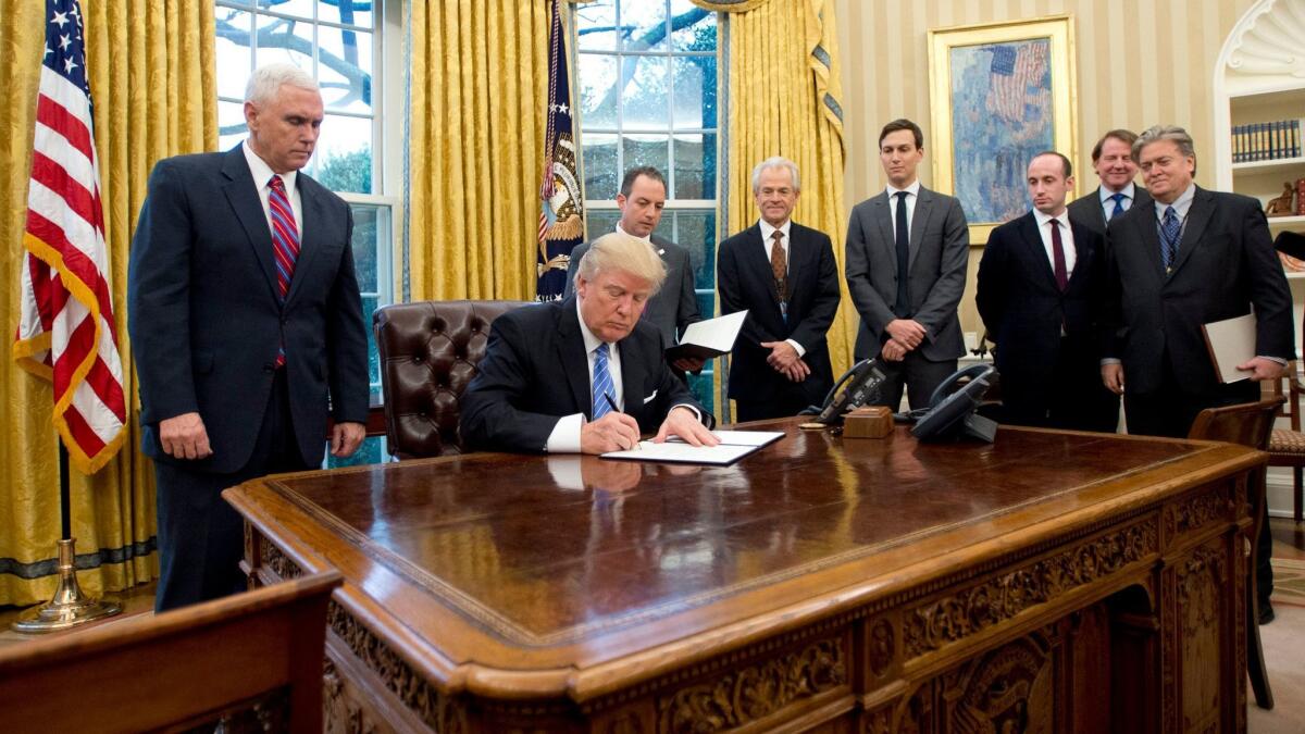 President Trump signs the first of three Executive Orders in the Oval Office of the White House on Jan 23, while his administration members and various advisers look on.
