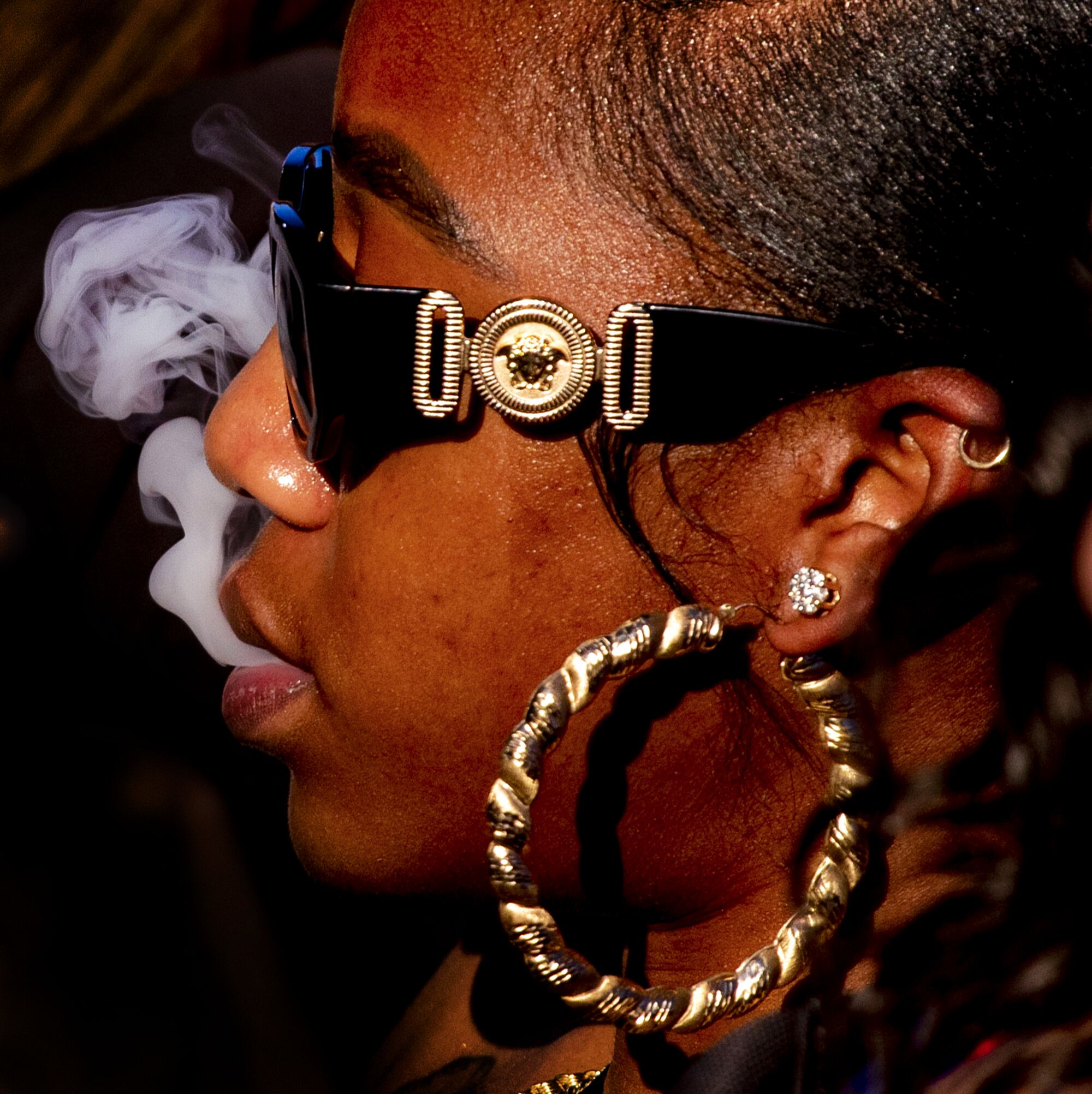 A woman wearing sunglasses and giant hoop earrings exhales smoke 