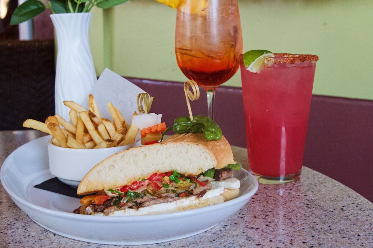A steak chimichurri sandwich and fries in foreground, a spritz, a hibiscus margarita and vase of fake flowers behind.
