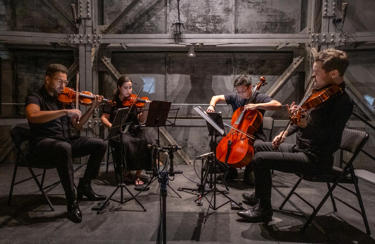 Four people playing in a string quartet in front of a metal industrial all.