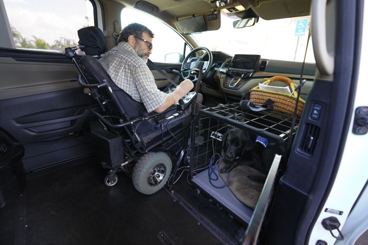 A man in a wheelchair sits behind the wheel of his modified vehicle, with a dog in a crate on the floor next to him. 