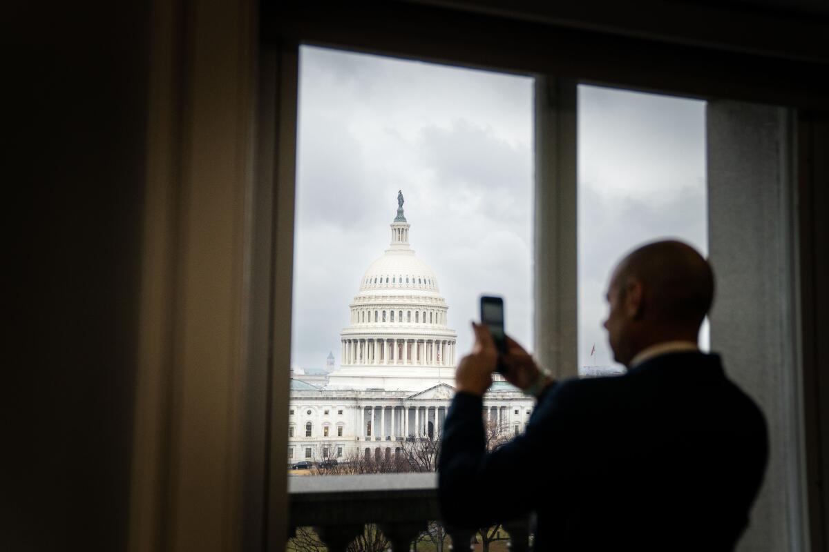 Standing near a window, a silhouetted man takes photograph of the outside, with the U.S. Capitol building in the center.