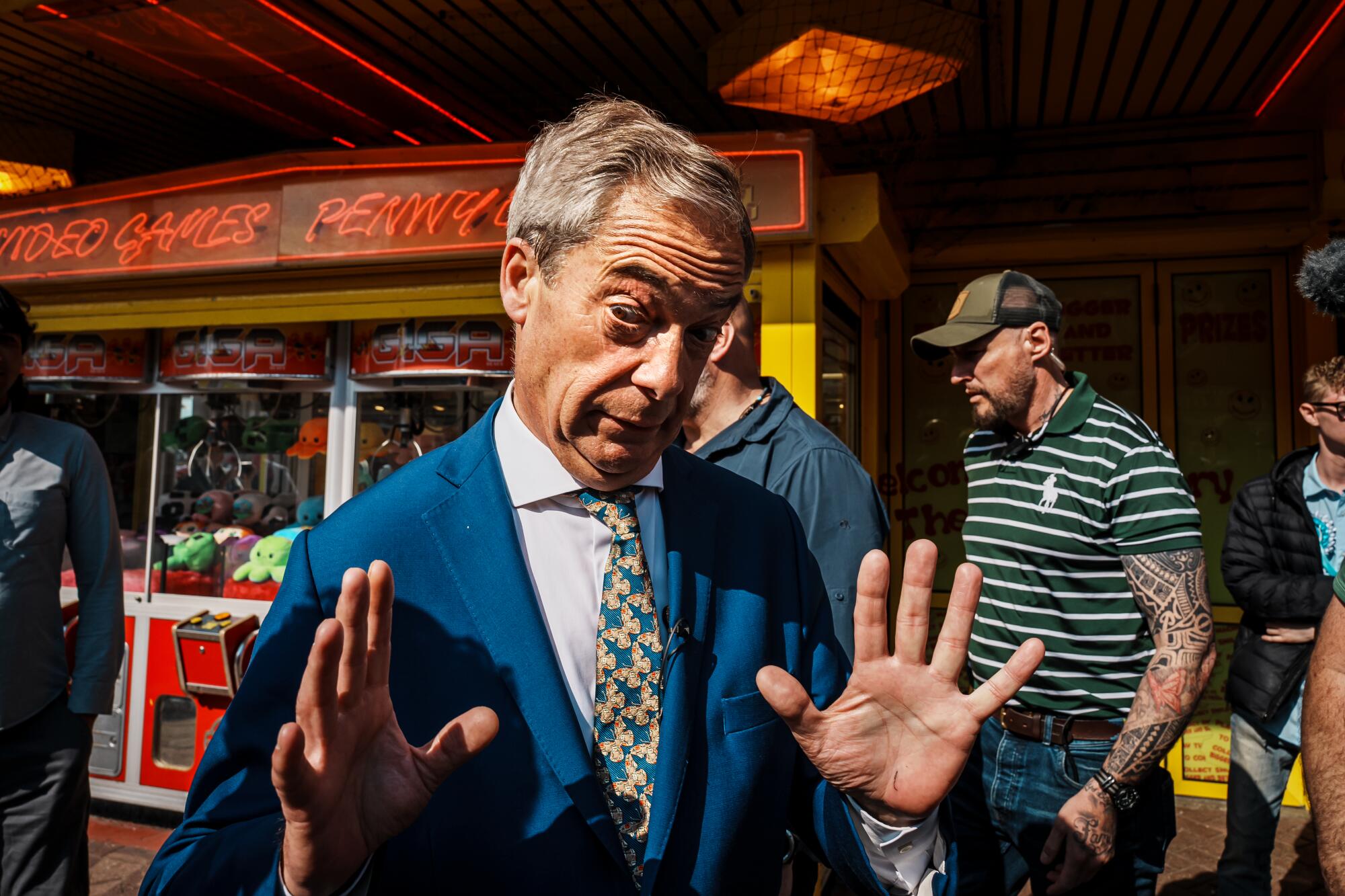 Nigel Farage raises his hands and looks toward the camera during a campaign event in an arcade. 
