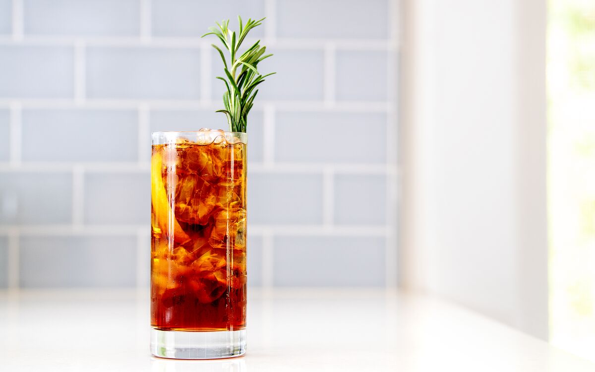 A tall glass is filled with ice and a brown beverage and a sprig of rosemary 