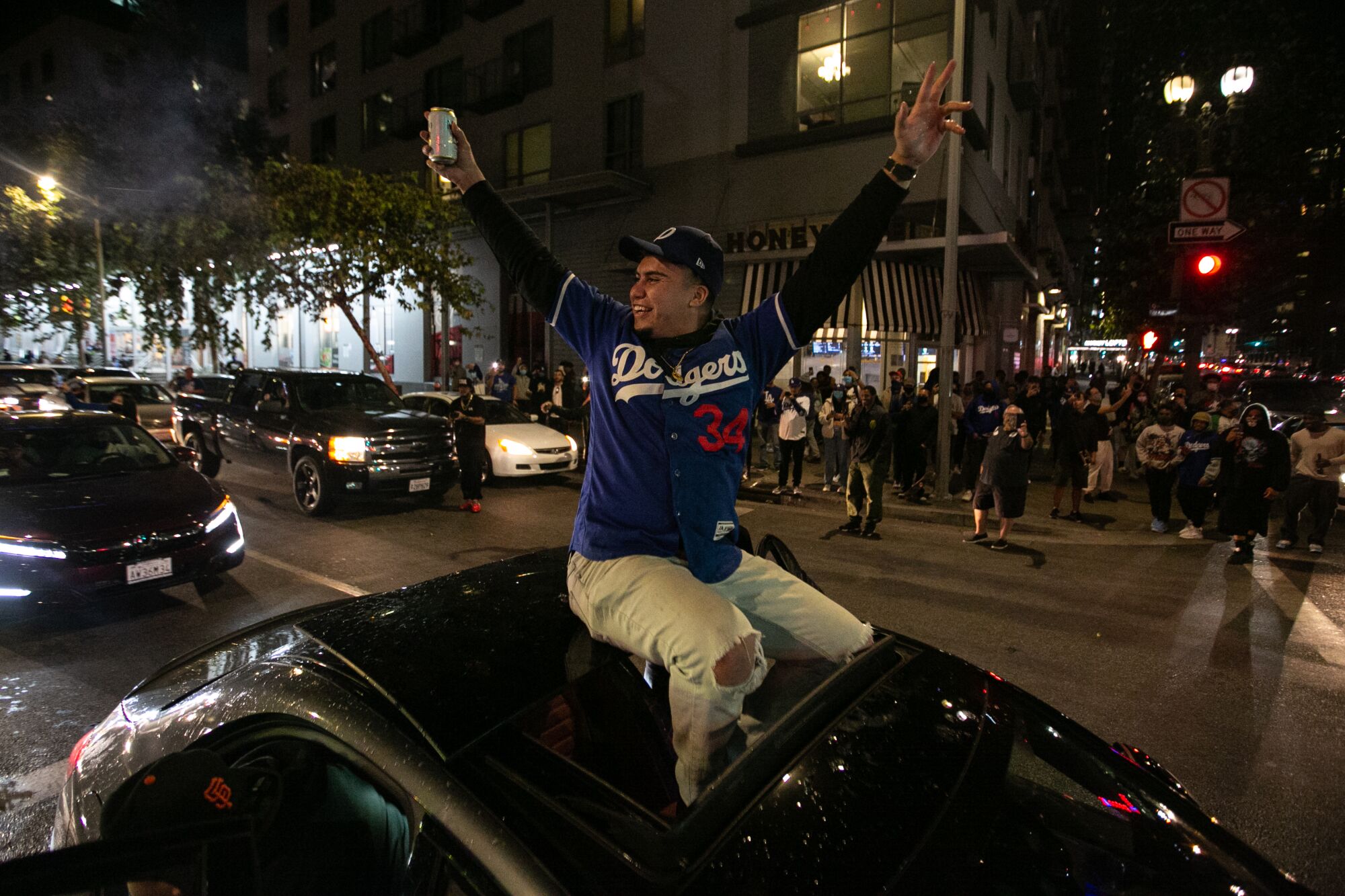 A Dodgers fan rides atop a moving car, arms raised, on a street.