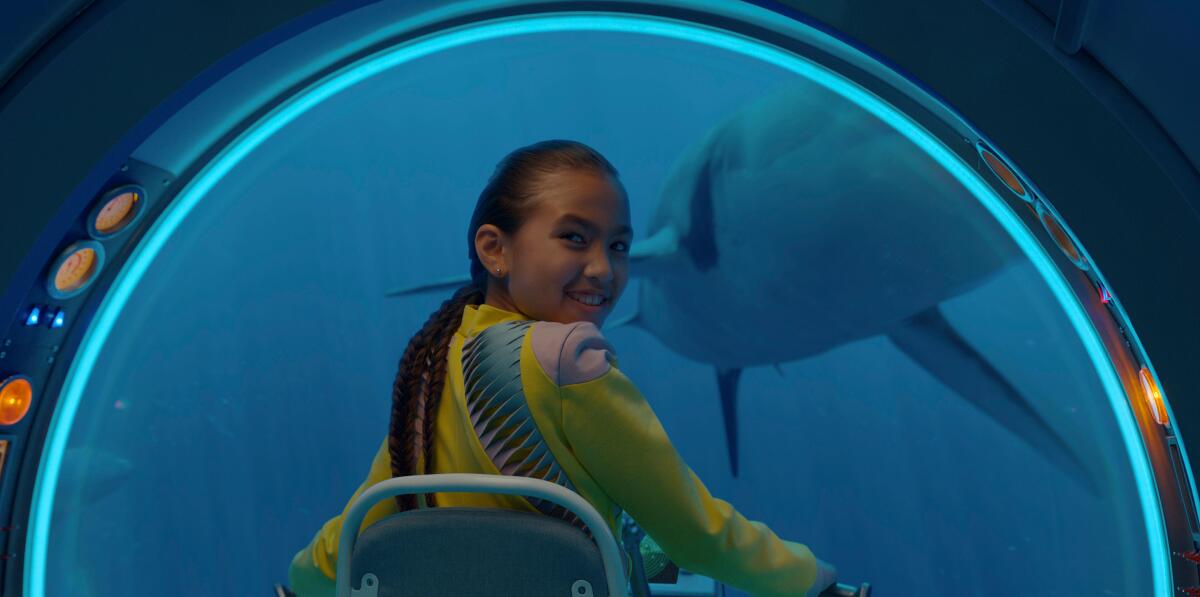 A young girl in a submarine, turning back to look at the camera as a shark passes outside the window.
