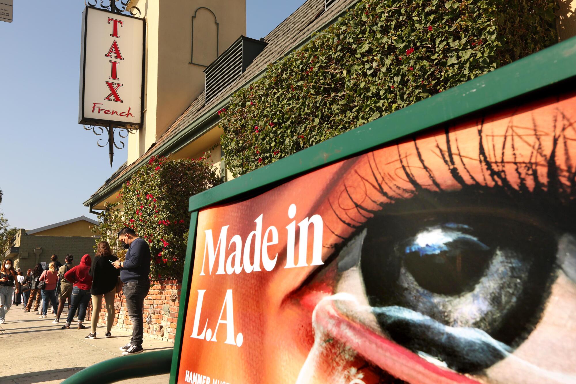 A line of people wait on a sidewalk outside a restaurant. In the foreground, a bus-bench ad says "Made in L.A."