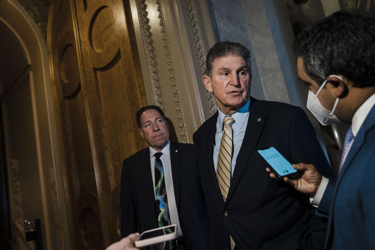 Sen. Joe Manchin stands in a hallway outside the Senate chamber and speaks with reporters holding phones.

