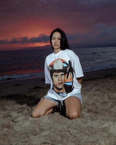 A person in a Paisa Boys T-shirt kneels on a sandy beach at sunset