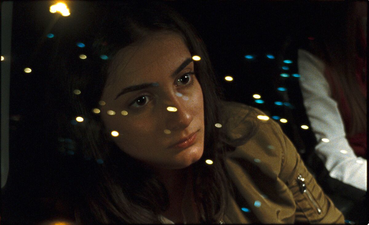 A young woman is seen in closeup through a car window; small lights reflect off the window.