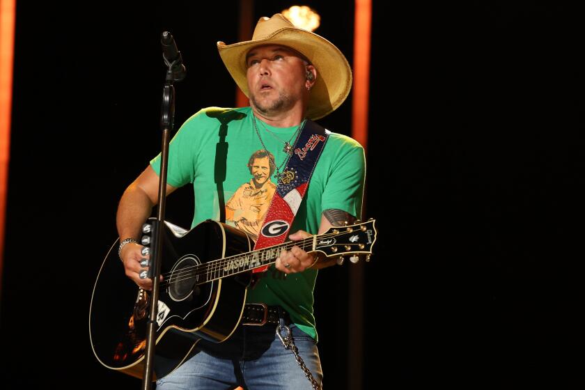 Jason Aldean is performing on stage, strumming a guitar and is wearing a green t shirt and and light brown cowboy hat