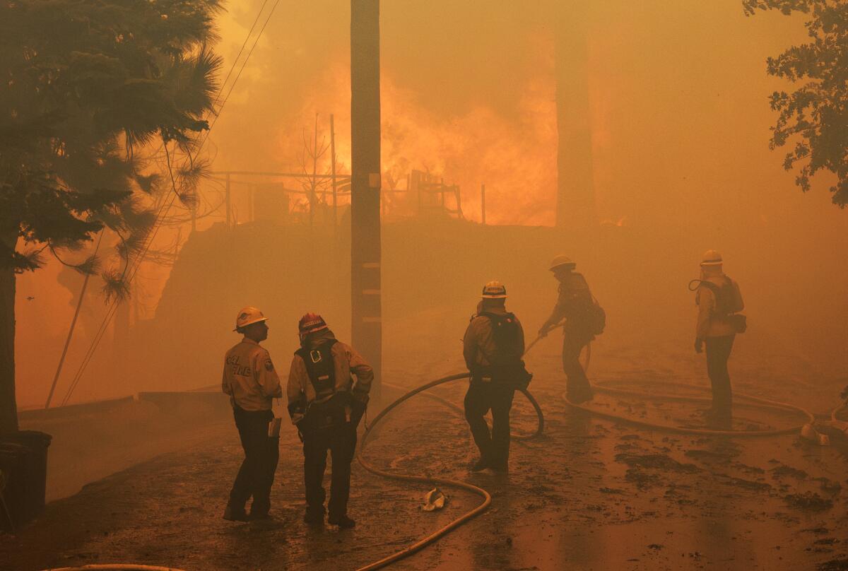 People, some in hard hats and holding a hose, stand in the orange haze from flames raging in the background