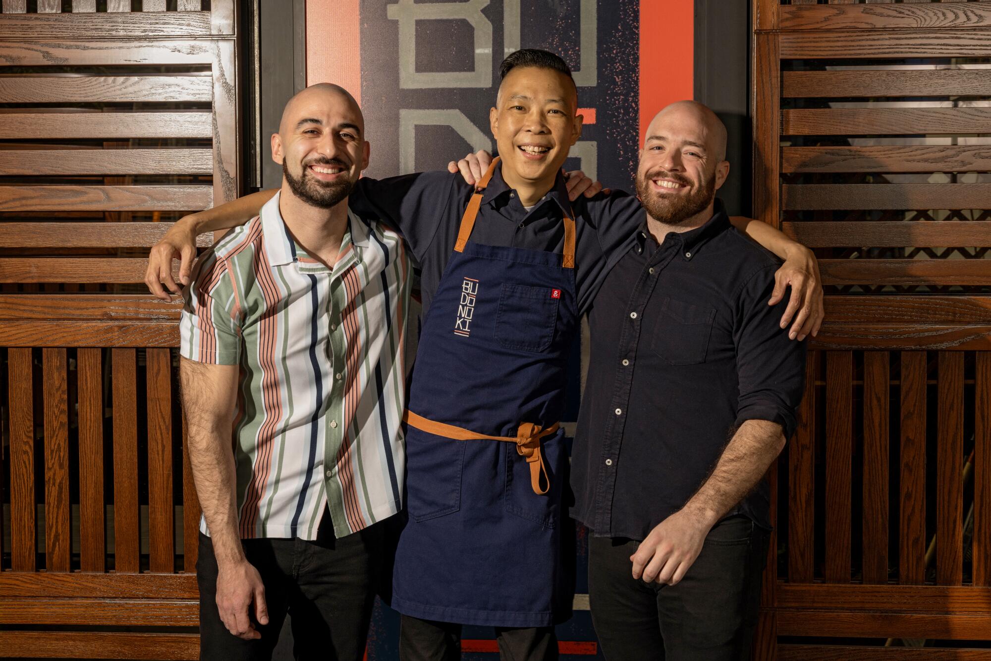 Three men stand smiling with their arms around one another's shoulders