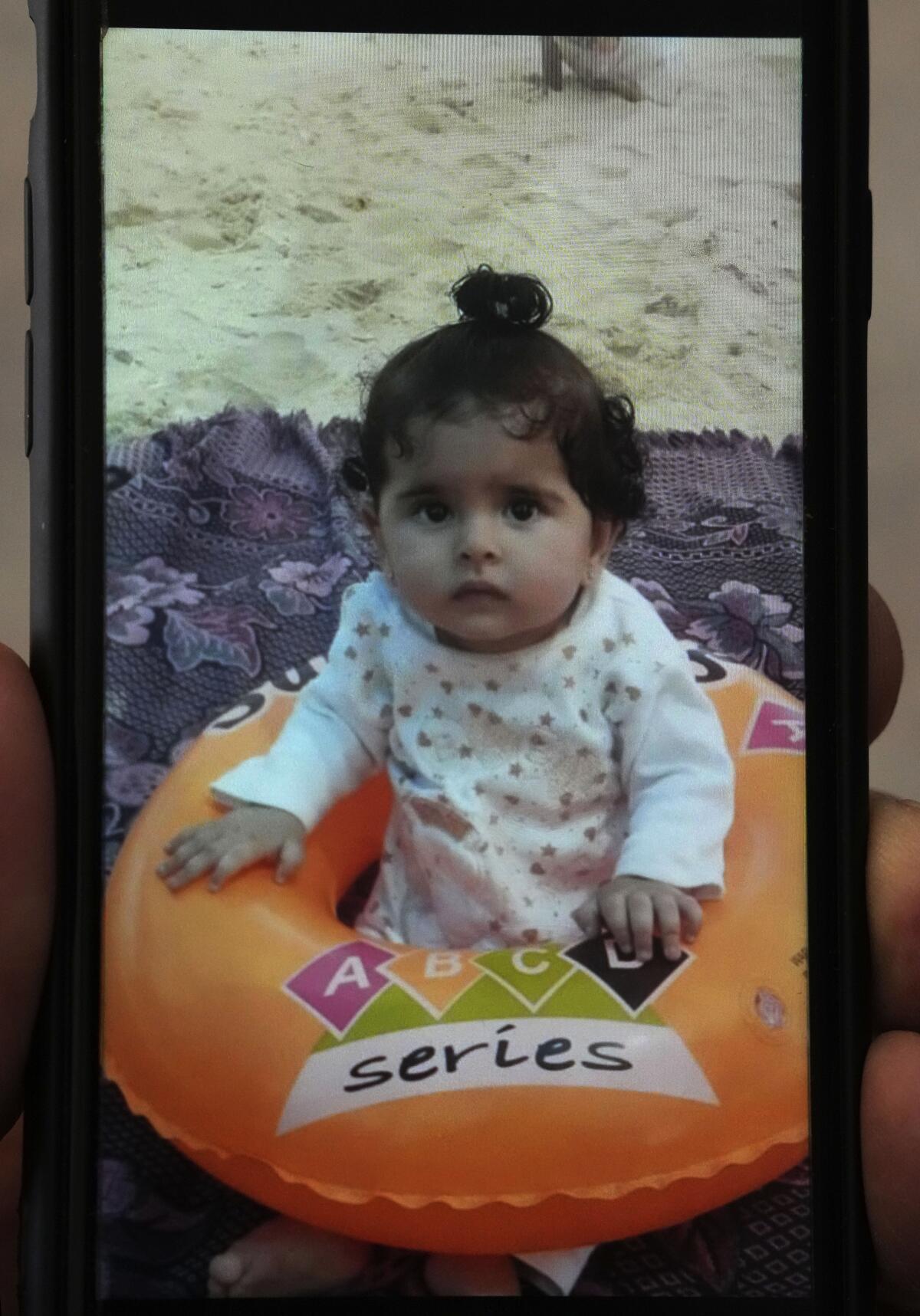 A phone showing a photo of a curly-haired baby girl sitting in an inflated toy on a rug