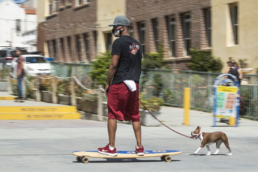 VENICE, CA-MAY 14, 2020: Ray Grey, 28, of Hollywood, wearing a mask to protect against the coronavirus,rmakes his way along the boardwalk in Venice Beach, on a long board with his dog Poet, a Boston Terrier running alongside. They are from Hollywood. A new rule, announced by Mayor Eric Garcetti on Wednesday, now requires face masks for all outdoor activities. (Mel Melcon/Los Angeles Times)