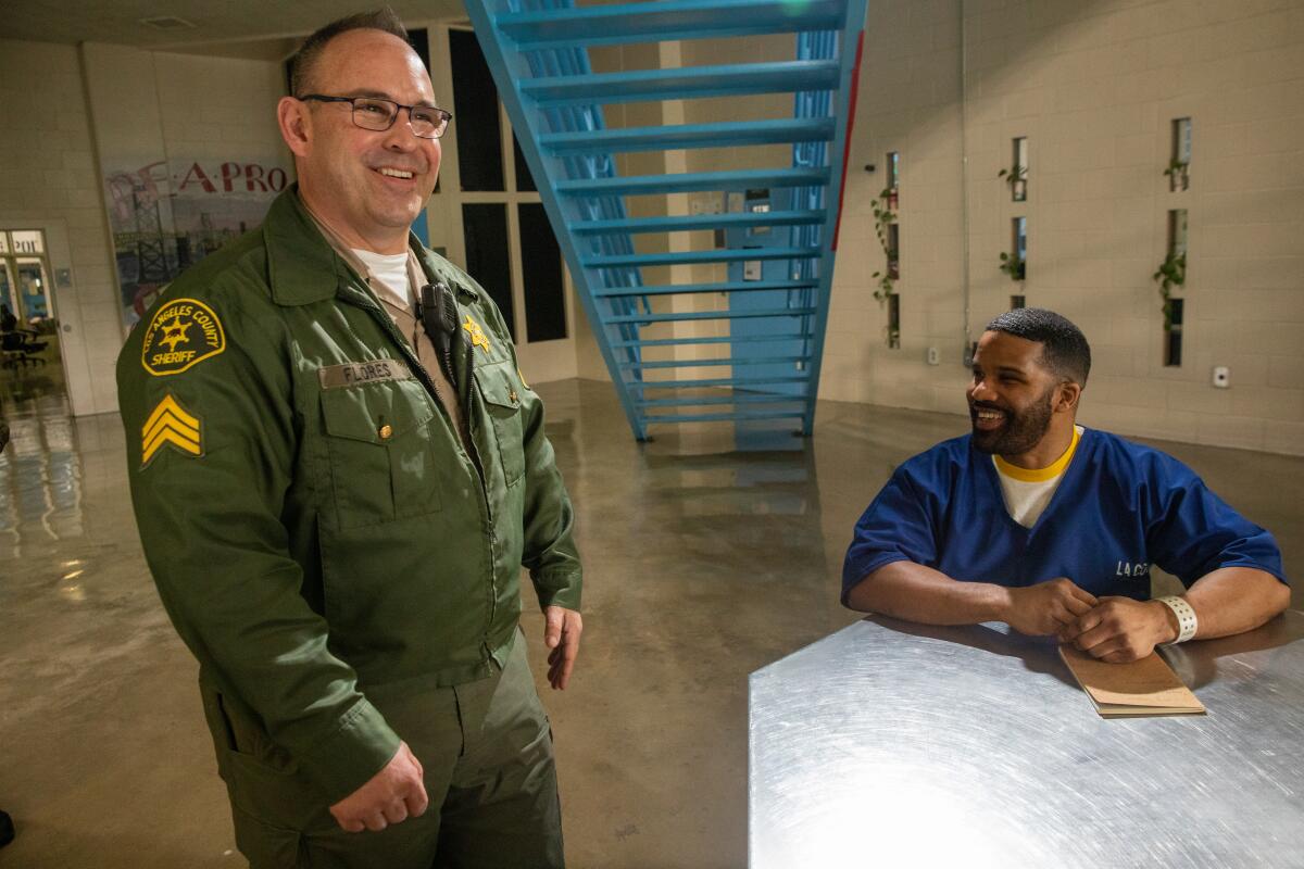 A man with glasses, in green uniform, smiles while standing next to a smiling man with a beard, in a blue top, who is seated 