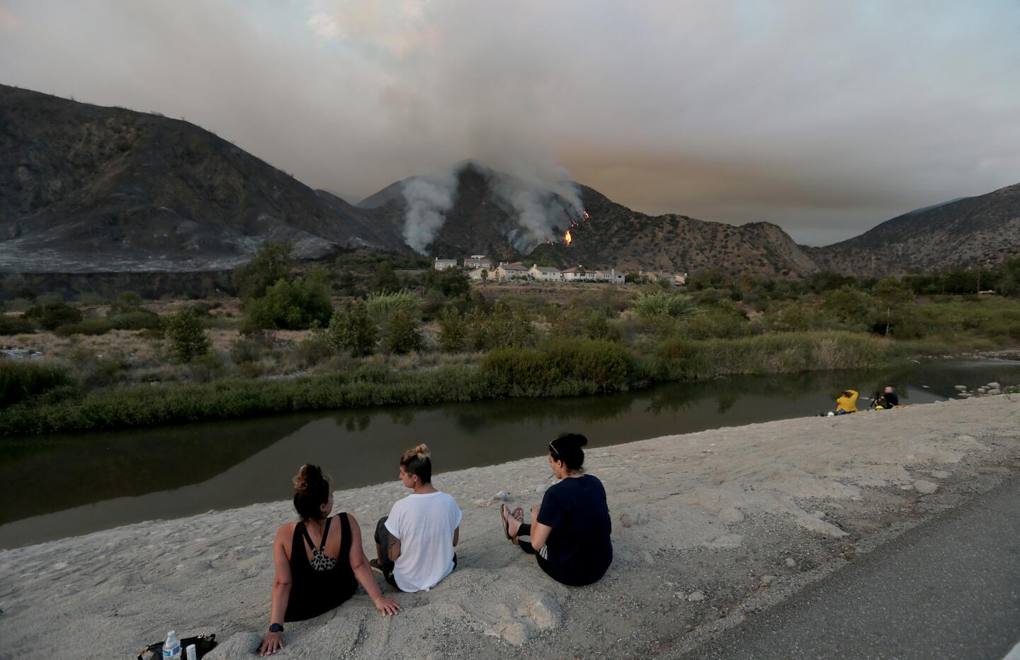 People sit on a riverbank and watch as the Ranch fire burns in the hills above a cluster of homes