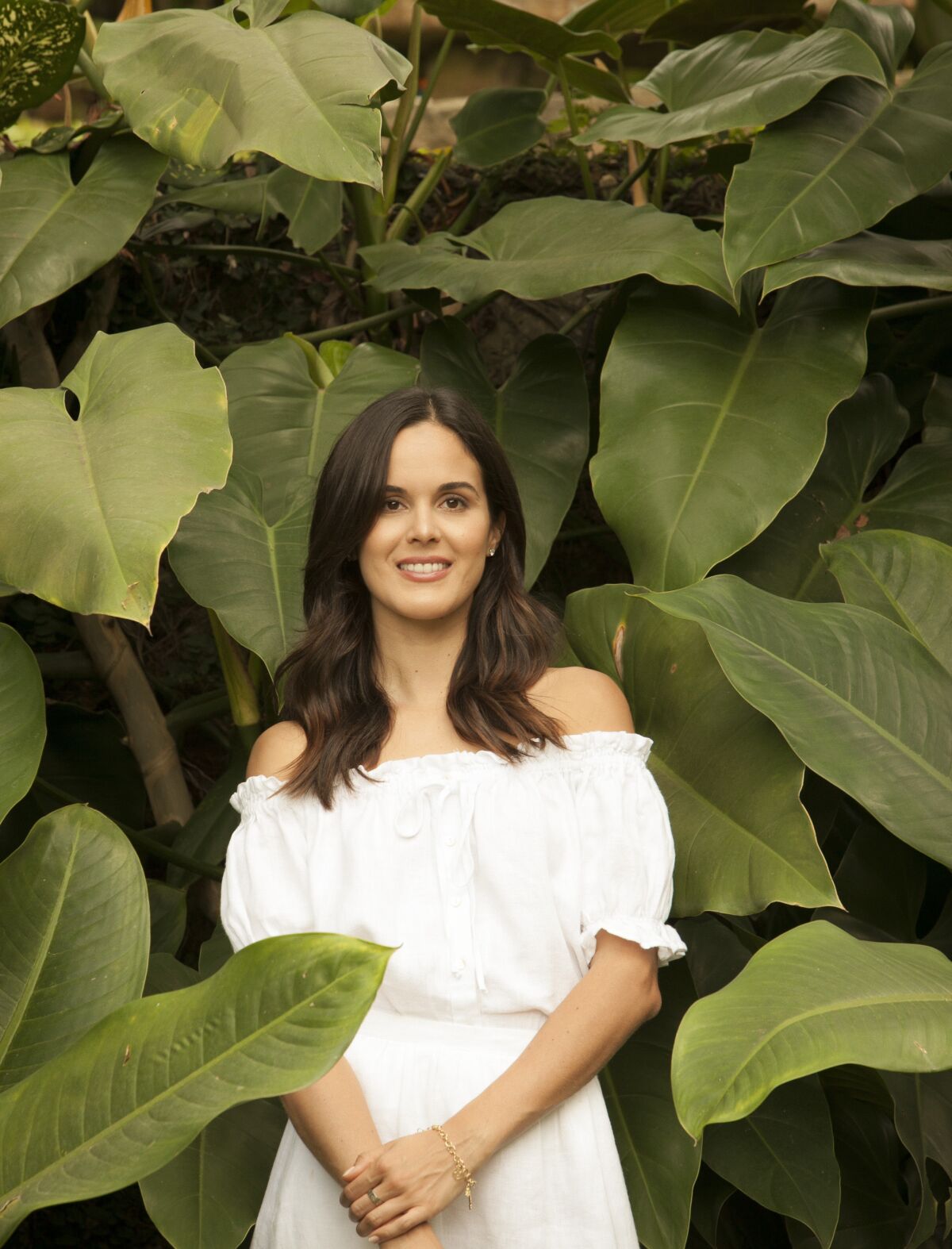 A woman in a white off-the shoulders dress stands surrounded by tropical foliage.