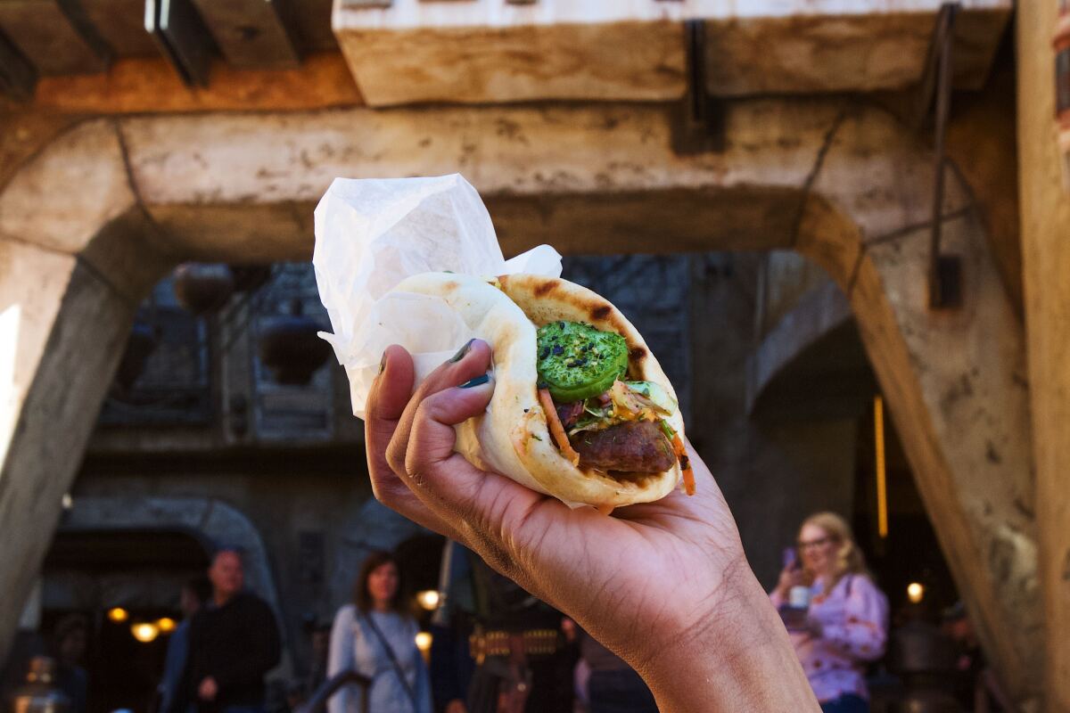 A hand holds a wrap bursting with Impossible sausage and slaw in front of an entrance to the Galaxy's Edge marketplace.