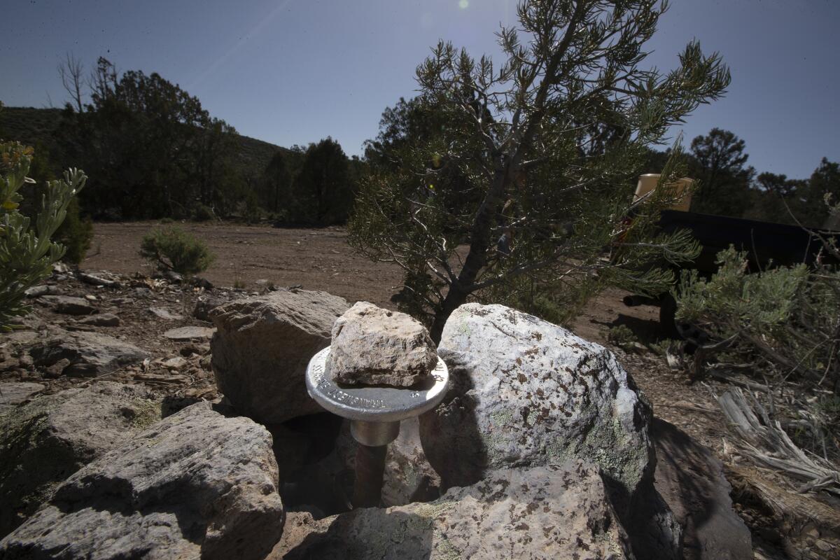 A rock sits on a silver pedestal among boulders next to trees and a dirt track.