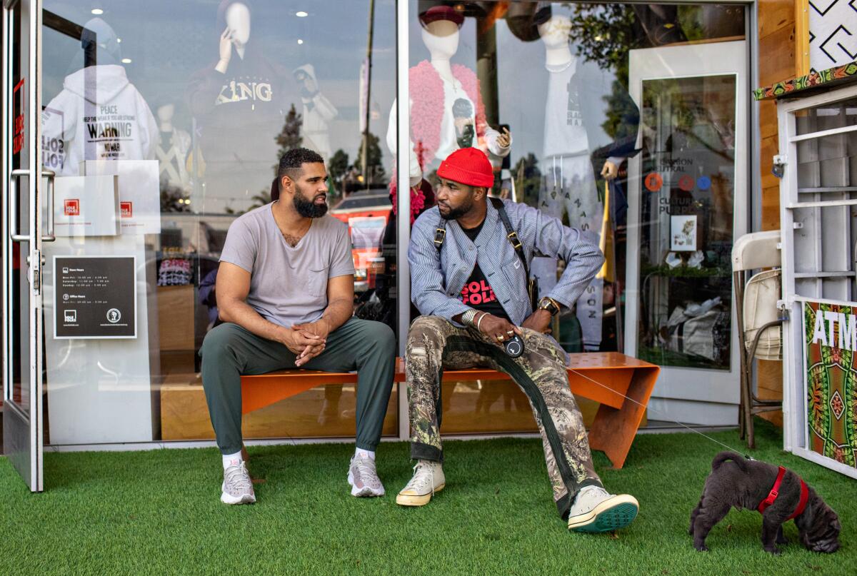 Two men sit on a bench in front of an apparel store; one has a dog on a leash.