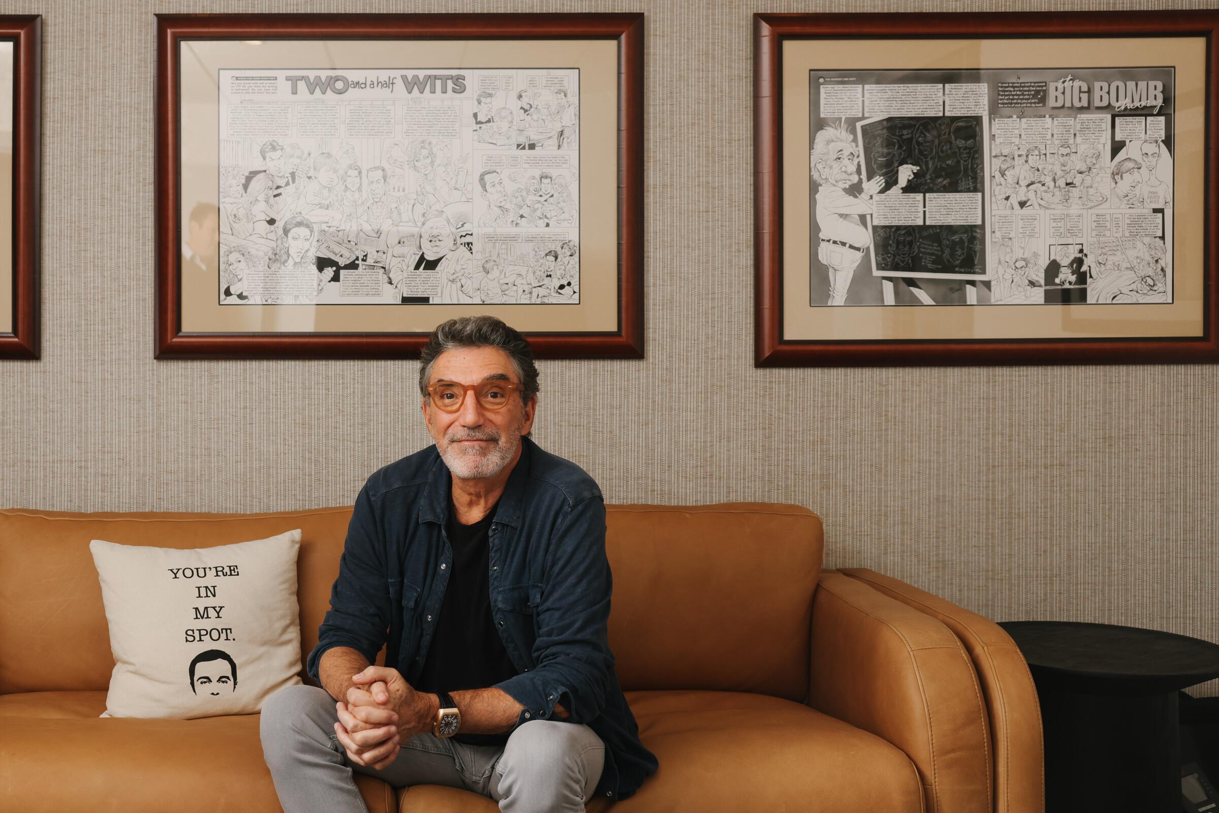 Chuck Lorre sits on a couch next to a white pillow that says "you are in my spot." Framed cartoon drawings hang on the wall.