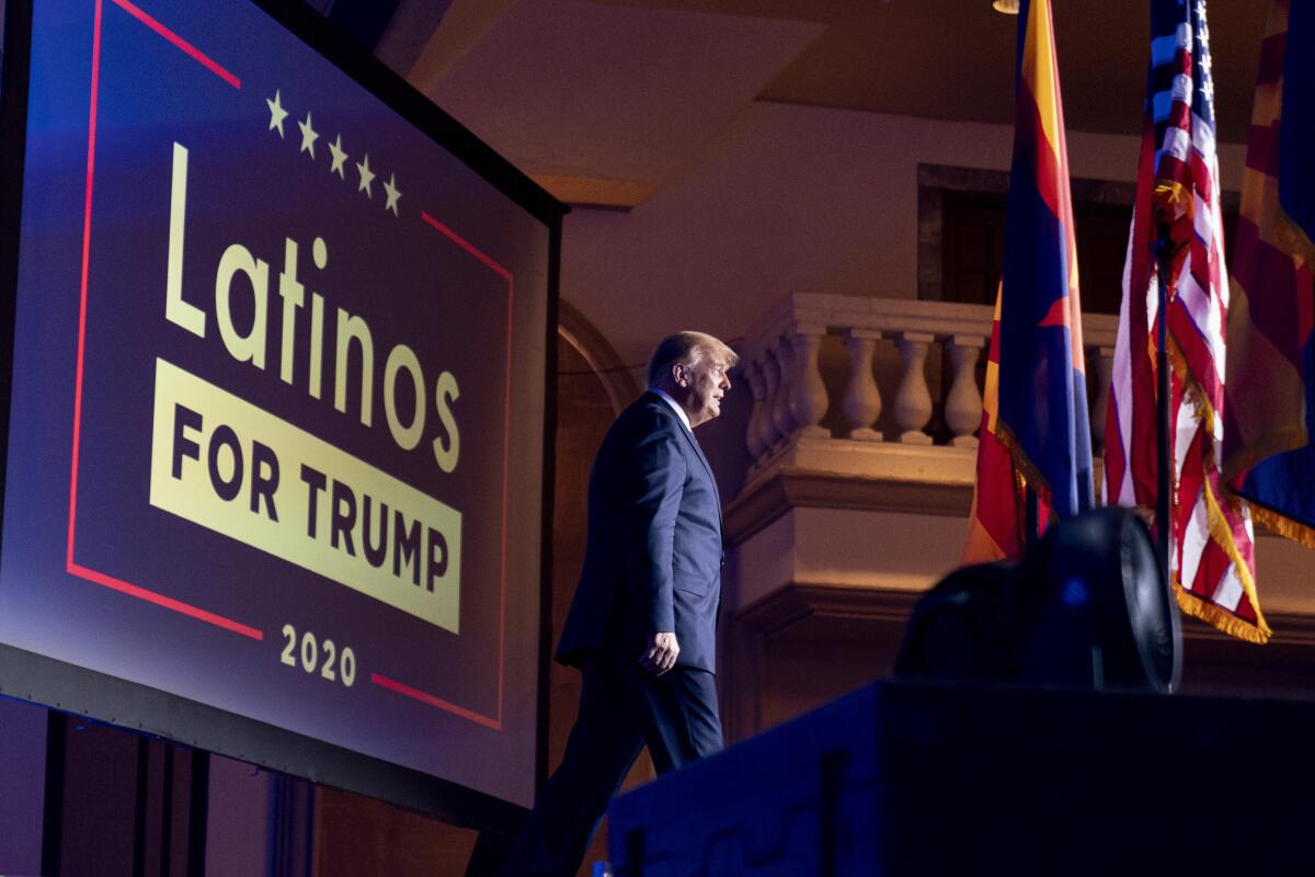 Donald Trump walks across a stage with a "Latinos for Trump 2020" sign behind him. 