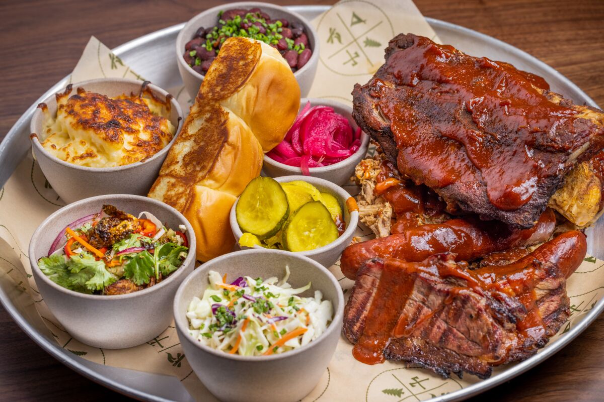 A circular metal barbecue tray holds ribs, tri-tip steak and pulled pork with pickles, mac and cheese and bread rolls.