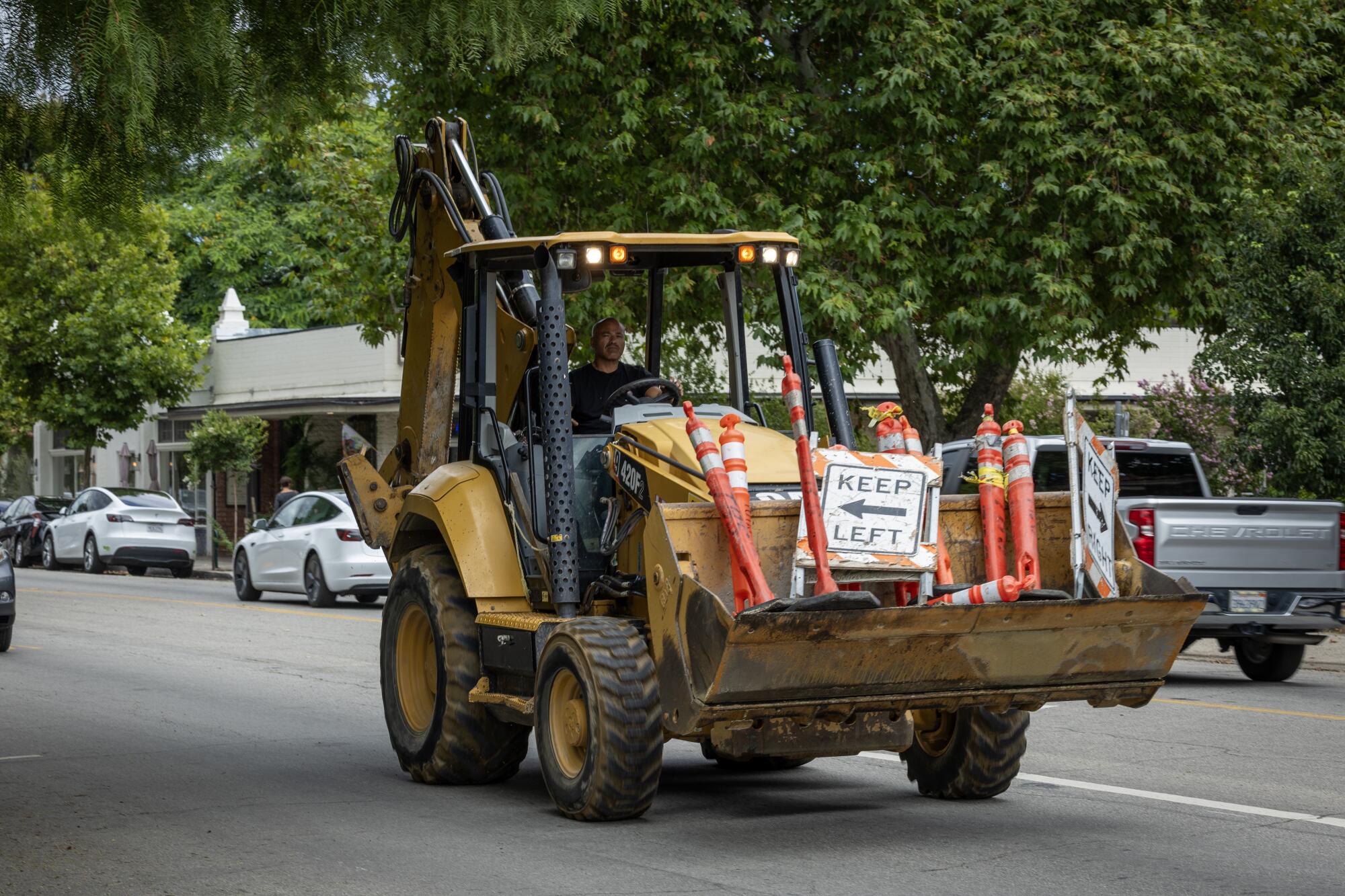 A backhoe loader carrying orange pylons and a keep-left sign drives down a tree-lined street.